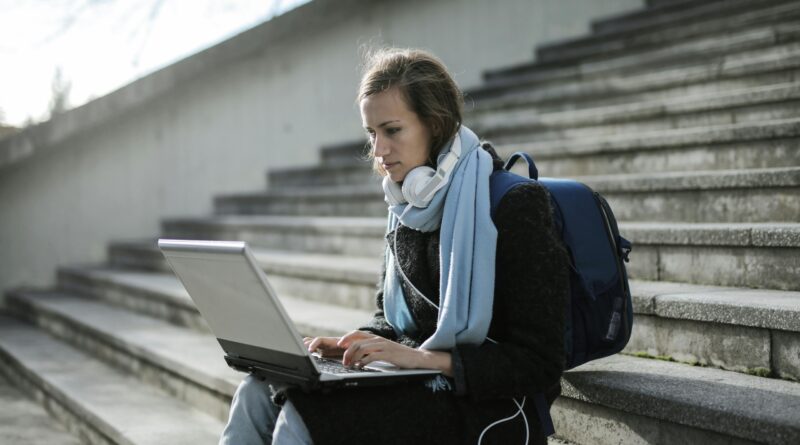 Woman using laptop on outdoor stairs, studying online with headphones and backpack.