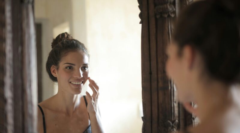 Young woman applying skincare and smiling at her reflection in a rustic mirror.